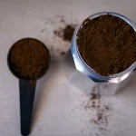 Imagem de pó de café em uma colher medidora é uma base de cafeteira moka onde se coloca o pó - estão sobre a mesa branca